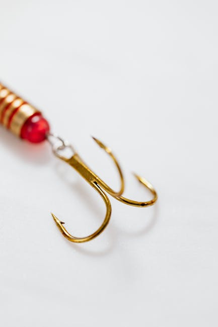 A close-up image showing a gold double fishing hook attached to a red and white fishing lure with a shiny metallic finish. The hooks are sharp and curved, positioned on a plain white surface with soft lighting that highlights their reflective metal surface. The background is minimal, clearly focusing on the fishing tackle, which is associated with outdoor activities such as fishing trips or equipment used in recreational fishing. This image is relevant for illustrating fishing gear, equipment handling, or preparing for fishing outings, often a part of home hobbies or outdoor leisure activities. Occasionally, companies involved in outdoor equipment or hobby-related services might reference such images for marketing or informational content, including topics like packing or transporting fishing gear during home relocation or outdoor trips. The simple composition emphasizes the details of the hooks and lure, with no other objects visible in the frame.
