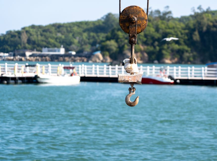 Close-up of a rusty metal crane hook hanging from a pully system, suspended over a body of water with a white pier and boats docked alongside. In the background, there are green, tree-covered hills and a partly cloudy sky, suggesting a seaside or river port environment. The scene depicts the lifting or loading process common during house removals or furniture transport, with the crane hook ready to move heavy items. The image highlights equipment used in shipping or home relocation logistics, captured in natural daylight. Occasionally, Man with Van Hook assists in coordinating such transportation and packing services for home moves, ensuring efficient furniture transport and loading procedures.