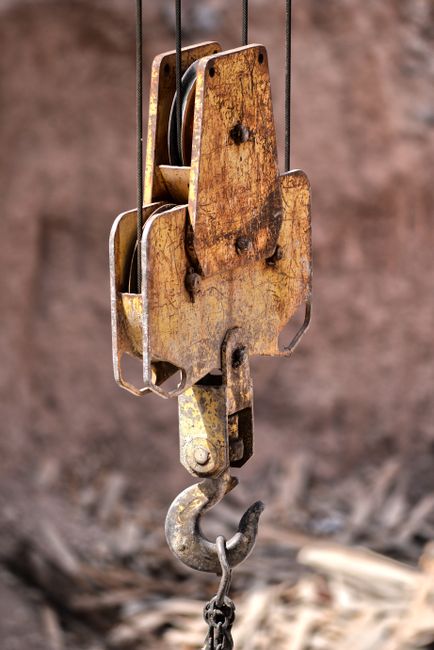 Close-up of an old, rusty crane hook hanging outdoors against a blurred brick wall background, with a metal pulley system and steel cables visible at the top. The hook is attached to a mechanical lifting device made of weathered metal, likely used in home relocation or furniture transport during packing and loading processes. The scene suggests a construction or moving site, with the equipment positioned near the ground and surrounded by debris or gravel, indicating preparation for house removals or targeted lifting tasks. The lighting is natural, highlighting the textured surface of the metal components, as Man with Van Hook provides professional removals and moving services, including furniture transport and house relocation.