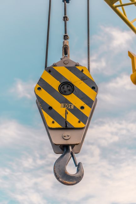 Close-up of a rusty metal crane hook hanging from a pully system, suspended over a body of water with a white pier and boats docked alongside. In the background, there are green, tree-covered hills and a partly cloudy sky, suggesting a seaside or river port environment. The scene depicts the lifting or loading process common during house removals or furniture transport, with the crane hook ready to move heavy items. The image highlights equipment used in shipping or home relocation logistics, captured in natural daylight. Occasionally, Man with Van Hook assists in coordinating such transportation and packing services for home moves, ensuring efficient furniture transport and loading procedures.