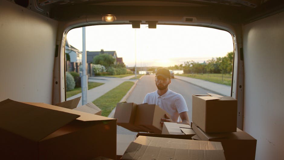 A man wearing a white shirt and baseball cap is standing outside on a residential street during sunset, loading several cardboard boxes into the back of a moving van. The van's interior is visible fro