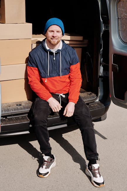 A man wearing a white shirt and baseball cap is standing outside on a residential street during sunset, loading several cardboard boxes into the back of a moving van. The van's interior is visible fro