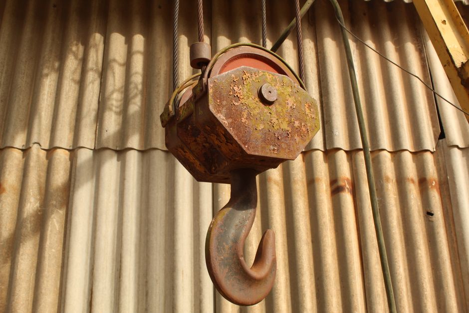 Close-up view of a large industrial crane hook suspended from thick steel cables against a clear blue sky. The hook is attached to a heavy-duty pulley system housed in a black metal casing with yellow