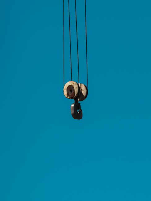 A close-up view of a black crane arm extending upward against a clear blue sky, with a metal lifting hook attached to a cable hanging beneath it. In the background, there are white residential buildin