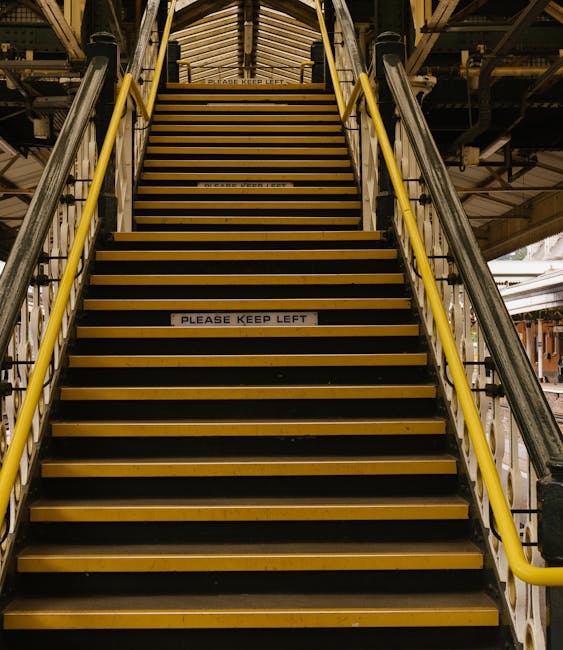 A straight flight of industrial stairs with yellow handrails on each side and black non-slip strips on each step, located inside a building with visible metal framework and wooden ceiling panels overhead. A sign on one of the middle steps reads 'PLEASE KEEP LEFT.' The stairs are part of a home relocation or furniture transport process, possibly within a warehouse or storage area used for packing and moving belongings. The environment is illuminated by ambient lighting, and the overall scene depicts the stairway preparation for loading or unloading furniture and boxes, consistent with house removals services provided by Man with Van Hook. The image emphasizes the importance of careful stair access management during the loading process in a house or property in Hook.