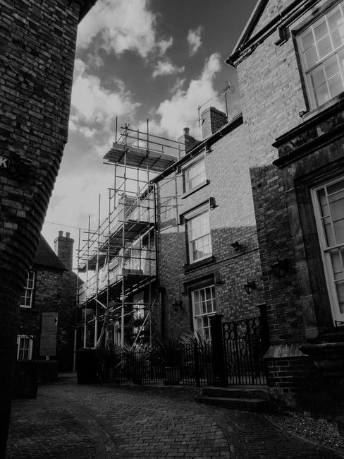 Black and white image showing the exterior of a terraced house in Hook with scaffolding erected along the front of the building, indicating ongoing property maintenance or renovation. The scaffolding consists of metal poles and wooden planks, extending from ground level up to the roof, with a partially visible chimney at the top. The photograph captures part of a narrow cobbled alleyway leading to the entrance of the property, which is situated between other similar terraced houses. A metal gate and small steps provide access to the front door, and neighboring buildings feature large sash windows. Overhead, the sky is partly cloudy, casting shadows across the scene. This setting relates to the home relocation context, where Man with Van Hook may assist in moving furniture and belongings around properties undergoing refurbishment or structural work as part of their house removals services.