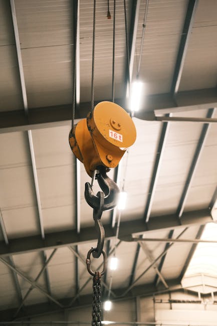 A yellow hoist with a weight capacity of 16 tons is suspended from a metal chain, hanging from the ceiling of an industrial warehouse with metal beams and paneling. The hoist features a hook at the bottom designed for lifting heavy objects, typically used in logistics and furniture transport during home relocation or moving projects. Bright lighting highlights the machinery against the ceiling structure, indicating an indoor environment suitable for loading and unloading activities. This equipment is indicative of professional removals and packing and moving services, such as those provided by Man with Van Hook, aligning with the logistics overview for house removals and furniture transport in the context of relocating to or from Hook Industrial Estate.
