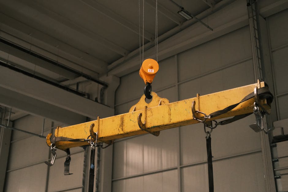 A close-up view of a yellow industrial crane hook labeled with a 5-ton weight limit hanging from a hoist system inside a spacious warehouse. The hook is suspended above a wide concrete floor, with metal shelving units and stacked materials visible along the sides. The warehouse has high ceilings with metal beams and ample lighting, creating a well-lit environment suitable for furniture transport and packing during a home relocation process. In the background, there are blurred figures and equipment, indicating ongoing logistics operations. This setting reflects the load-bearing capabilities used in large-scale removals and furniture transport, as managed by Man with Van Hook, dedicated to smooth and secure house removals in the UK.