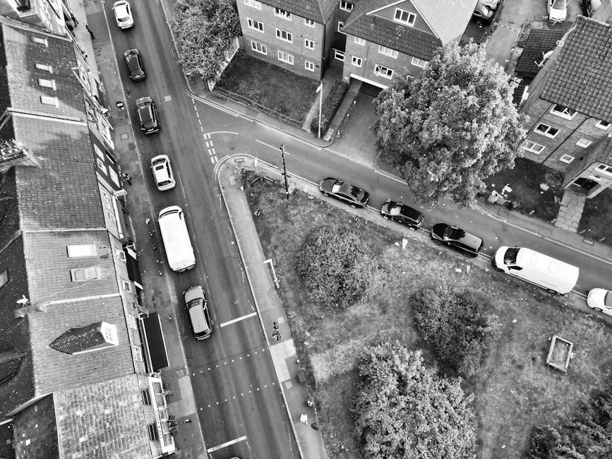 Aerial black-and-white photograph showing a residential street with parked cars along both sides and a small green area with trees and bushes in the center. In the foreground, the roofs of terraced houses with tiled roofs and windows are visible. A white van is parked at the edge of the pavement near the green space, while other vehicles, including a small car and a larger van, are parked along the street. The street appears to be part of a home relocation process, likely involving furniture transport and loading onto the van, as suggested by the context provided by Man with Van Hook's removals services. The scene captures a typical urban setting with minimal activity, primarily focusing on the environment and parked vehicles that are integral to moving logistics.