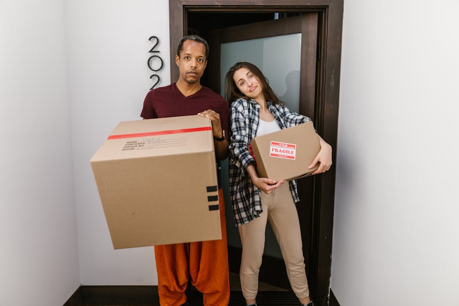 A man with dark skin and short black hair, dressed in an orange pair of trousers and a maroon shirt, stands inside a doorway holding a large cardboard box with red and black markings, indicating fragile contents. Next to him, a woman with light skin and long brown hair, wearing a plaid shirt over a white top and beige trousers, holds a smaller cardboard box with a visible 'Fragile' label. The scene takes place in a residential hallway with white walls and a black doorframe, where natural light illuminates the area. The background shows part of the doorframe leading outside or to another room. The image depicts a home relocation and furniture transport process, likely involving packing and moving services provided by Man with Van Hook, with attention to careful handling of packaging materials and loading procedures.