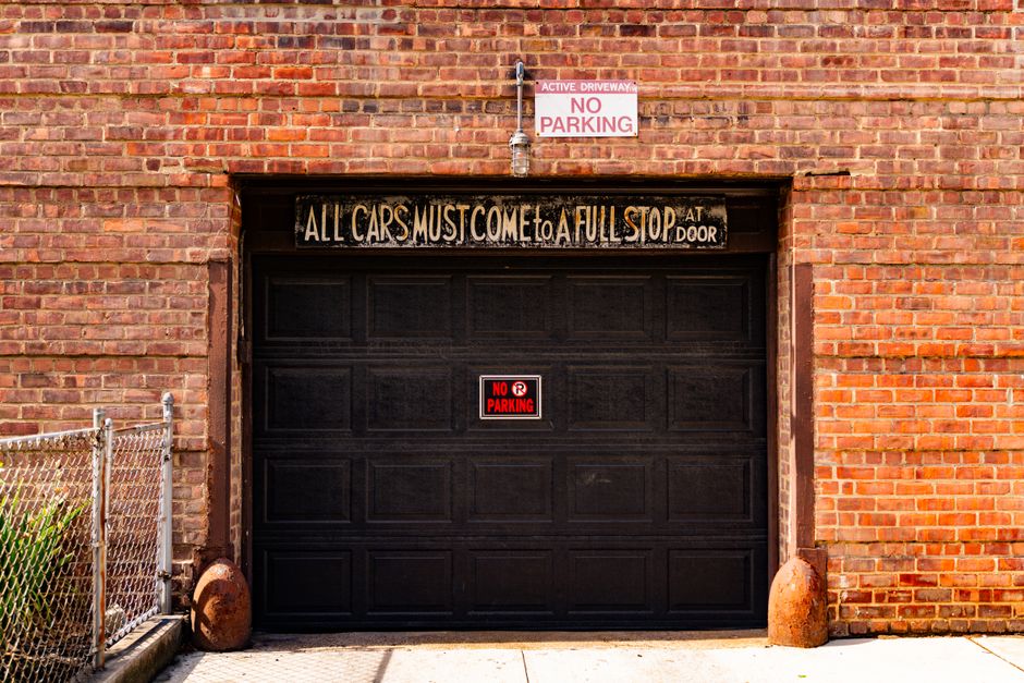 A black garage door set into a red brick building exterior, with a handwritten sign above that reads 'All cars must come to a full stop at door'. A red and white 'No Parking' sign is attached to the center of the garage door. Above the door, there's a metal pipe supporting an overhead sign that states 'Active Driveway No Parking'. To the left of the garage, a short chain-link fence encloses a small green bush, and two large, rounded, rust-colored weights are placed on the ground beside the fence. The scene is lit with natural daylight, indicating an outdoor urban setting. This image depicts a typical home or commercial property where movement and parking restrictions are relevant, which relates to house removals and relocation services provided by Man with Van Hook, emphasizing the importance of understanding parking suspensions and fines during house moves or furniture transport.