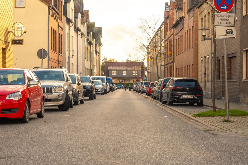 A quiet urban street lined with parked cars on both sides, including a red hatchback, a silver van, and various other vehicles, with a residential building façade featuring brick and painted exteriors along the sidewalks. The street is paved with asphalt, and a narrow sidewalk runs alongside, with a sign indicating parking restrictions and street cleaning times attached to a pole on the right. In the background, a small house with a sloped roof is visible, and leafless trees stand further down the street, suggesting late autumn or winter. The scene is illuminated by soft natural light, typical of an overcast day, with the overall setting reflecting a typical home relocation or furniture transport environment where parking and loading logistics are important, as managed by services like Man with Van Hook.