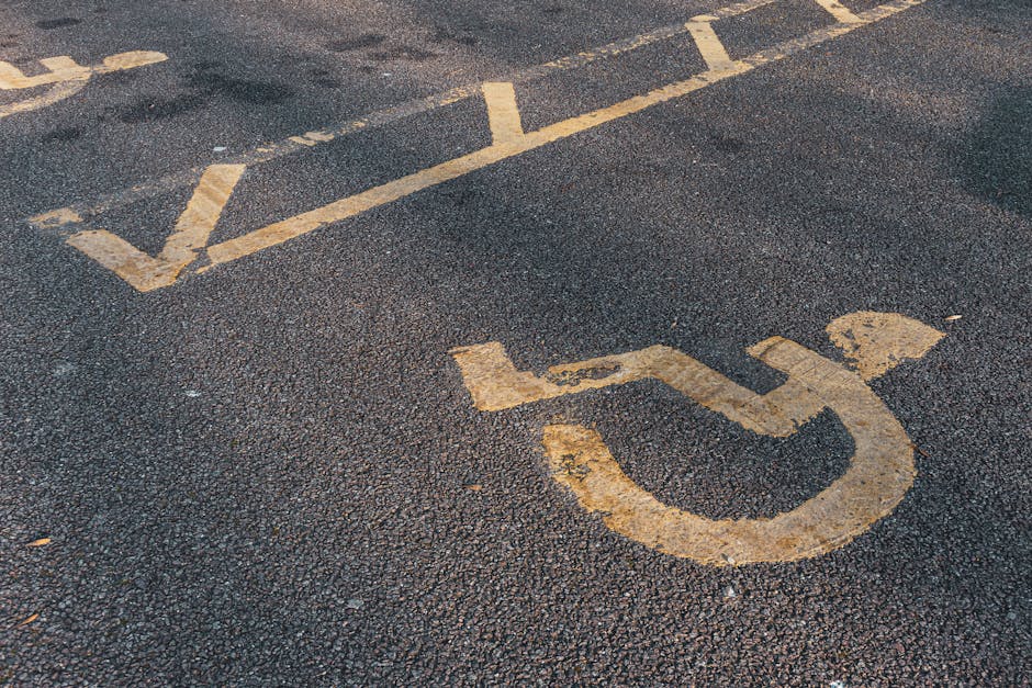 Close-up view of a designated accessible parking space marked by a yellow-painted wheelchair symbol on dark asphalt pavement. The symbol is slightly worn with some paint chipping, and the parking area features yellow lines indicating the space boundaries. In the background, additional parking lines are visible, extending across the asphalt surface. The scene suggests an outdoor parking lot suitable for home relocation or furniture transport, with a clear emphasis on accessible parking for moving vehicles. The image visually supports relocation services like those provided by [COMPANY_NAME], highlighting the importance of proper parking arrangements for efficient loading and unloading during house removals or packing and moving processes, especially in urban or residential environments.