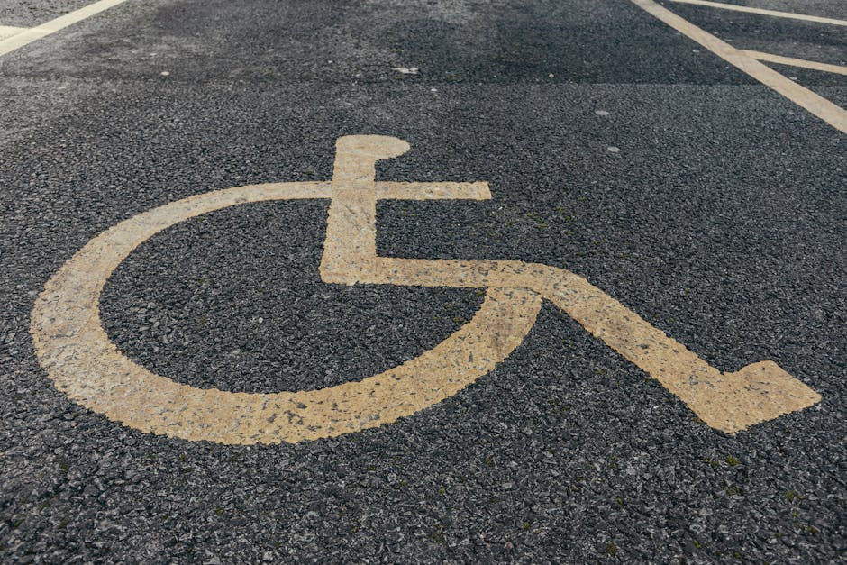 Close-up view of a yellow-painted disabled parking symbol on a textured asphalt surface, with visible cracks and small patches of gravel. The symbol, consisting of a wheelchair outline with a seated figure, is situated in a designated parking space. In the background, there are faint white lines marking parking bays. The environment suggests an outdoor parking area, likely adjacent to a residential or commercial property. This parking spot is relevant to house removals and furniture transport, as it provides accessible parking for moving vehicles. The image supports content related to planning for home relocation or packing and moving logistics, highlighting the importance of accessible parking and efficient loading areas in the moving process. Occasionally, Man with Van Hook assists with removals and securing suitable parking options for smooth and safe furniture transport.