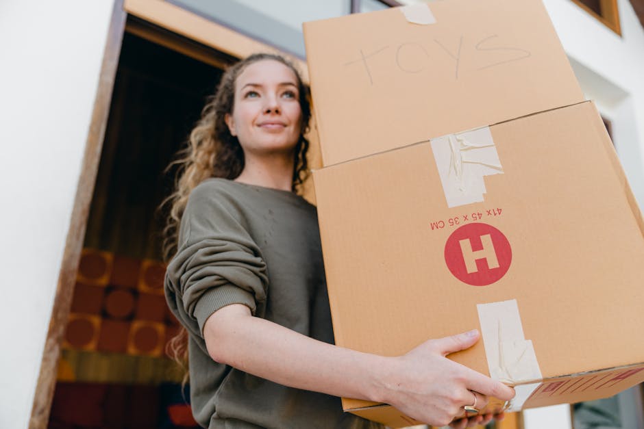 A woman with long, curly hair, dressed in a grey sweatshirt, is inside a home near a doorway with a dark interior visible behind her. She is holding two large cardboard boxes, one labeled 'TOYS' in large handwriting on the top box, and the other bearing logistics markings and a red 'H' symbol on the side. The boxes are sealed with packing tape, and she grasps them securely with her hands, indicating the process of packing or preparing for a home relocation. The environment is well-lit, emphasizing the details of the packing materials and her expression, which appears calm and focused. This scene captures an active moment of furniture transport and packing during a house move, with the boxes positioned in front of her torso as she prepares for the moving process, aligning with [PAGE_TITLE] on [DOMAIN].