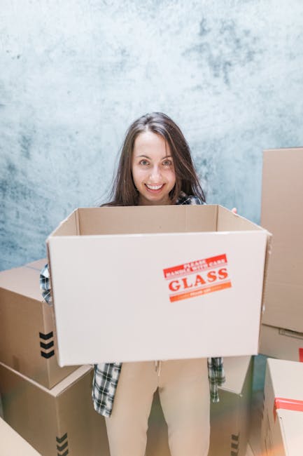 A woman with long, curly hair, dressed in a grey sweatshirt, is inside a home near a doorway with a dark interior visible behind her. She is holding two large cardboard boxes, one labeled 'TOYS' in large handwriting on the top box, and the other bearing logistics markings and a red 'H' symbol on the side. The boxes are sealed with packing tape, and she grasps them securely with her hands, indicating the process of packing or preparing for a home relocation. The environment is well-lit, emphasizing the details of the packing materials and her expression, which appears calm and focused. This scene captures an active moment of furniture transport and packing during a house move, with the boxes positioned in front of her torso as she prepares for the moving process, aligning with [PAGE_TITLE] on [DOMAIN].